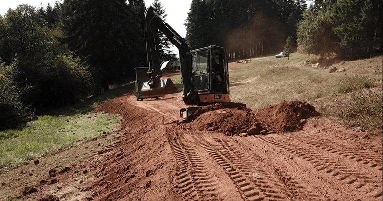 Small excavator on hillside clearing soil with forested background and mounded earth visible