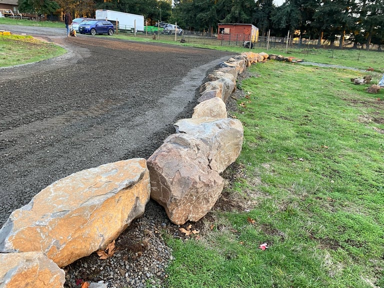 Large boulders lining a dirt driveway on a rural property with parked vehicles and buildings in the background
