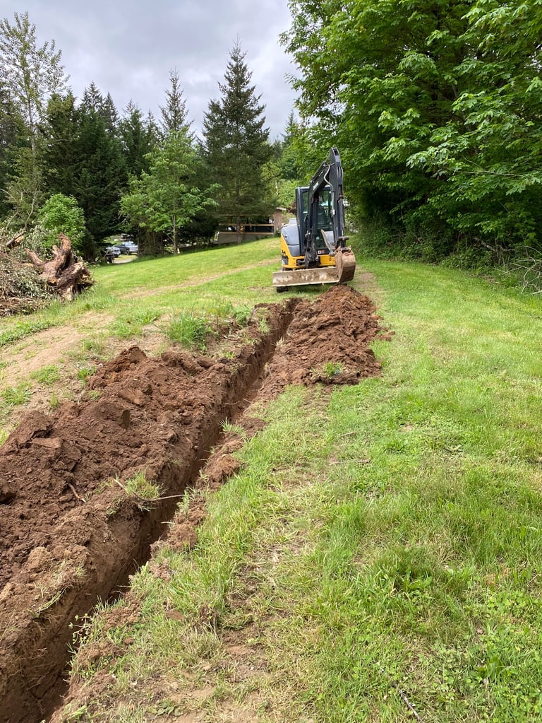 Mini excavator digging a trench in a grassy field with trees in the background