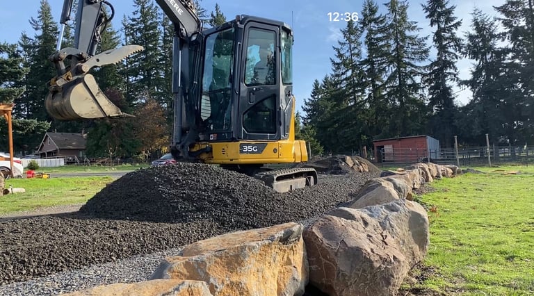 Yellow and black excavator digging gravel with piles of rocks and logs on a grassy property surrounded by evergreen trees