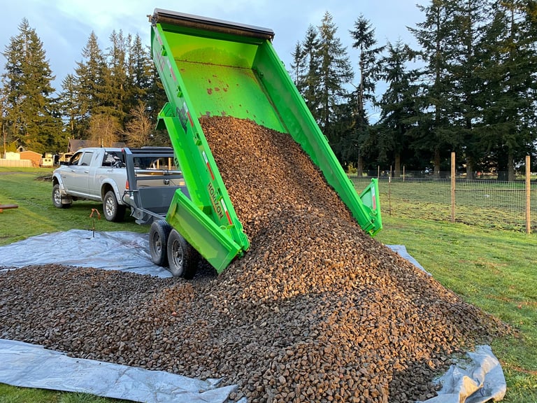 Bright green dump truck unloading gravel onto white fabric in rural setting with evergreen trees and white pickup truck in background