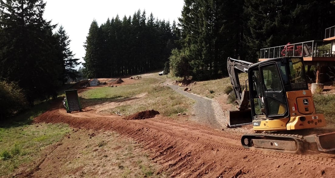 Small yellow and orange excavator operating on a dirt construction site surrounded by coniferous trees