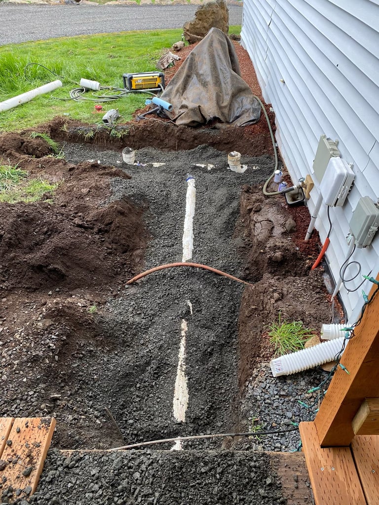 Drainage trench dug along the side of a house with white PVC pipes laid in gravel, near completed landscaping work