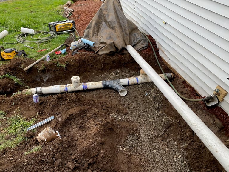 Excavated drainage and plumbing pipes laid in a trench beside a house with white siding during foundation work