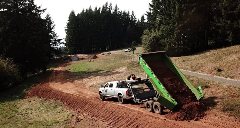 White pickup truck with bright green dump trailer unloading mulch onto dirt ground surrounded by evergreen trees