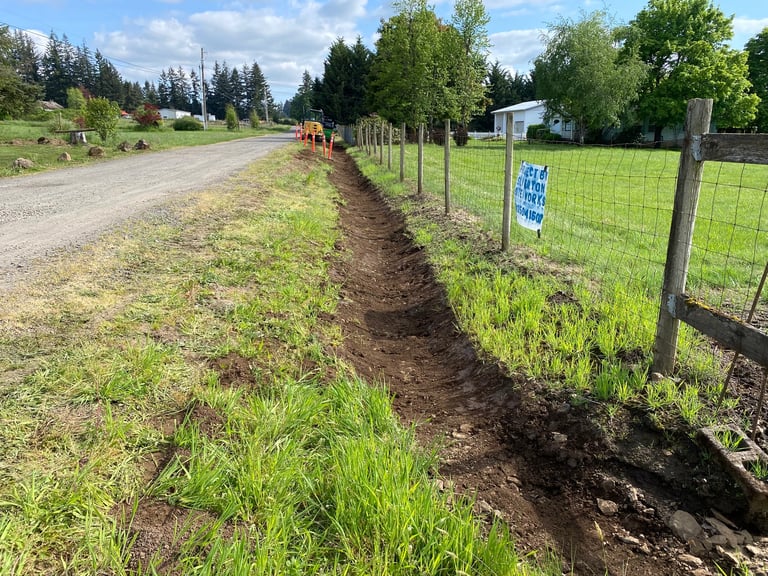 Rural property with gravel driveway, metal fencing, grassy field, and coniferous trees in background on sunny day