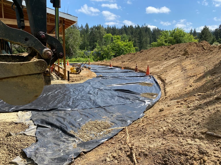 Construction site with black landscape fabric laid on dirt, heavy equipment visible, coniferous forest in background