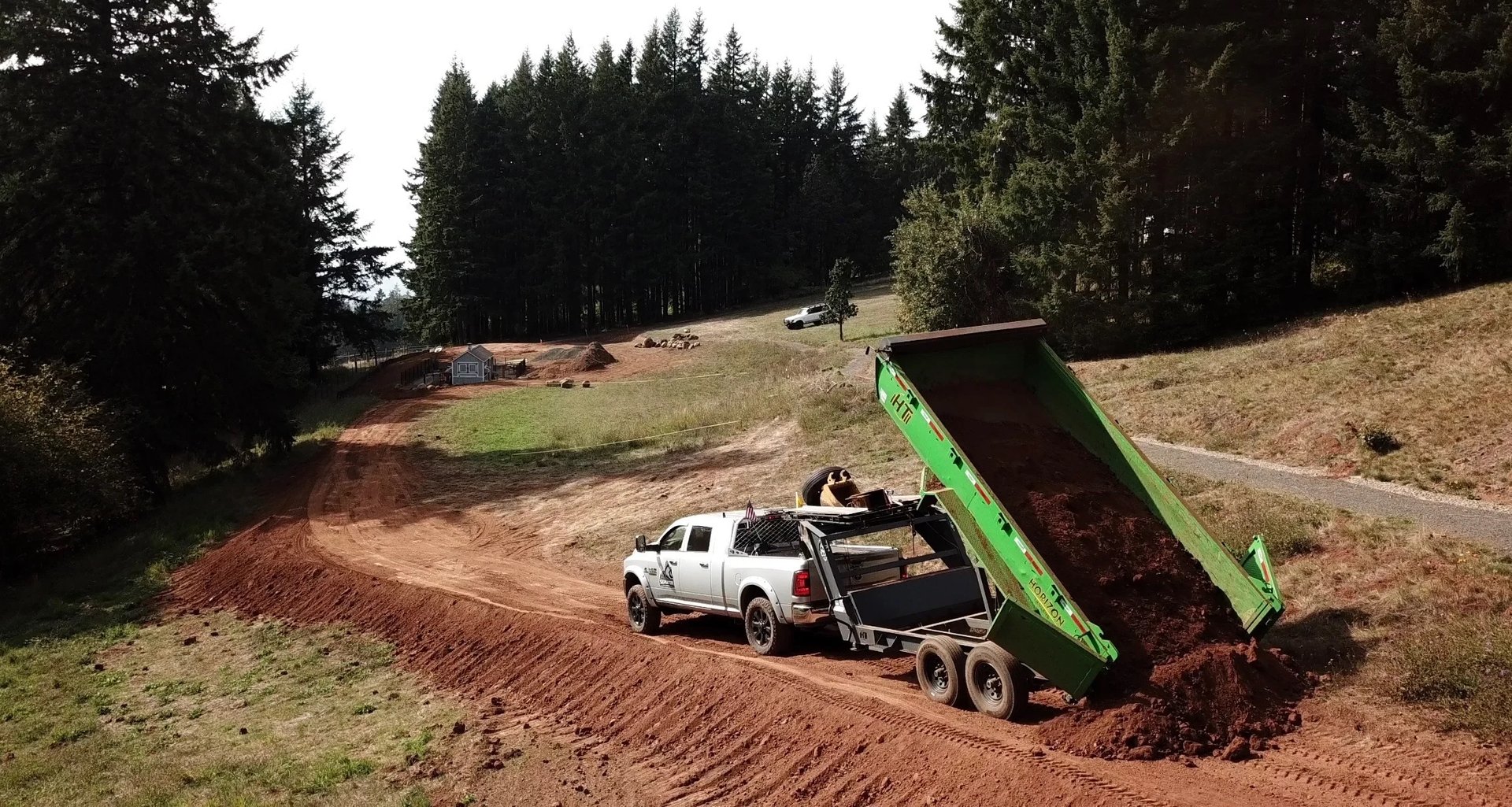 White pickup truck with green dump trailer unloading dirt onto a construction site surrounded by evergreen trees