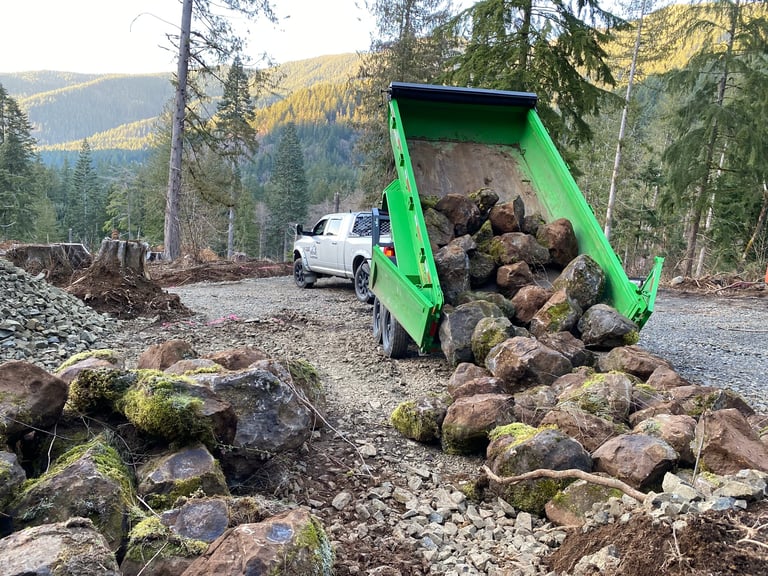 Green dump truck unloading rocks and logs in a forested mountain area with a white vehicle nearby