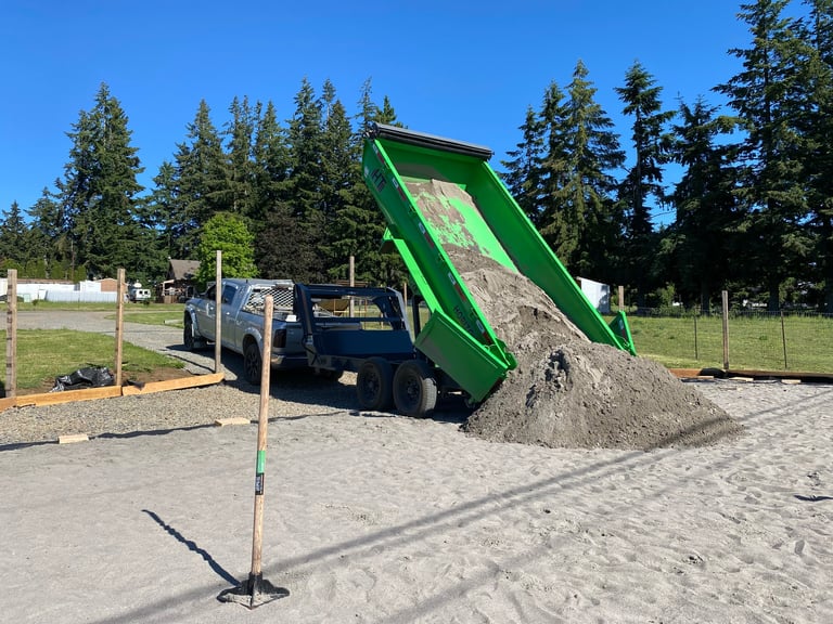Dump truck with green raised bed unloading gravel onto sandy ground with evergreen trees and park in background