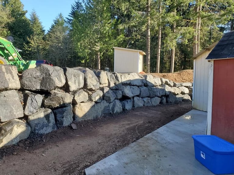 Large stacked boulders forming a retaining wall along a property with coniferous trees and utility buildings in background