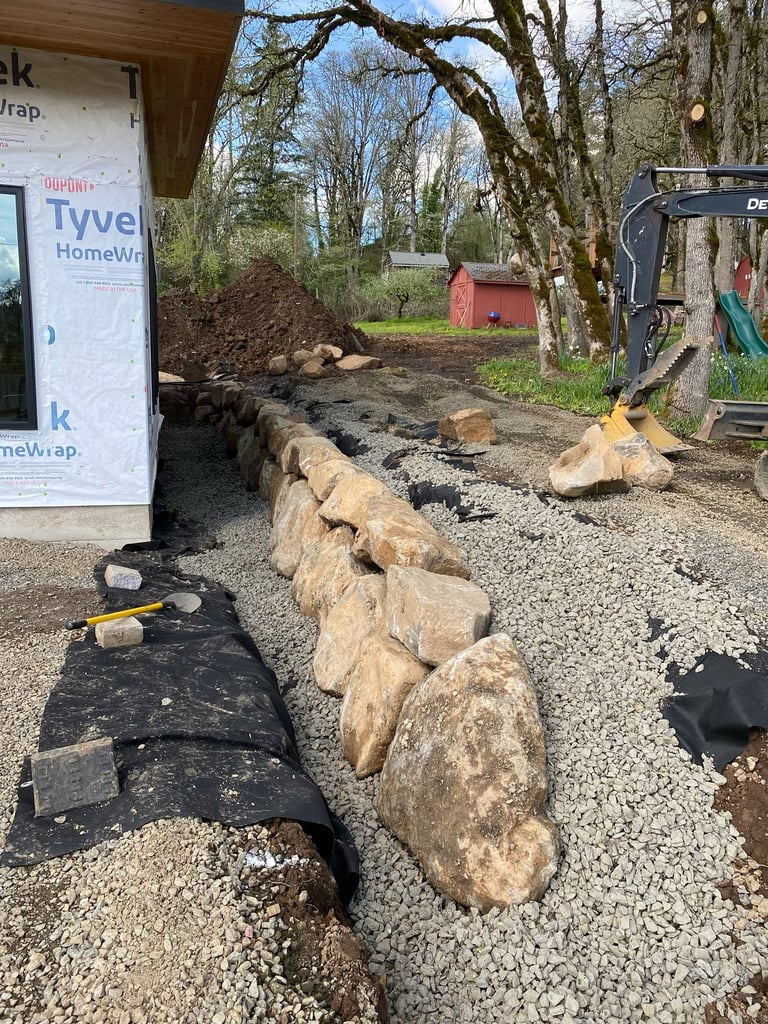 Landscaping work site with large stones arranged in a line, excavator equipment, bare trees, and a red shed in background