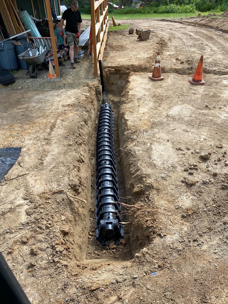 Black drainage pipe laid in a trench during residential construction, with orange cones and a building structure visible in the background