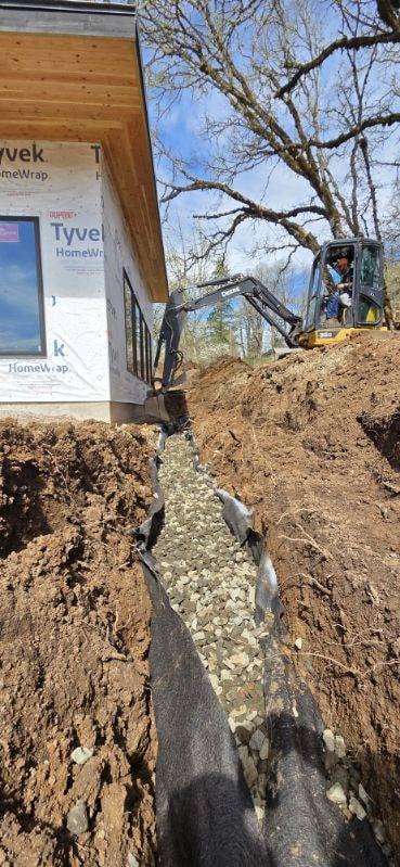 Construction site with mini excavator trenching near wooden structure with Tyvek wrap and bare trees