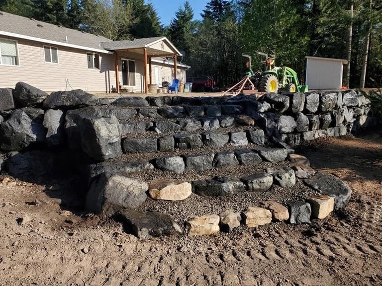 Construction crew assembling a large retaining wall made of stacked boulders in front of a residential home surrounded by evergreen trees