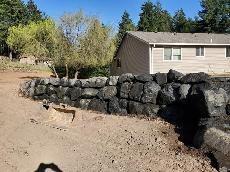 Rural property with a stacked rock wall, dirt yard, and beige house surrounded by pine and birch trees