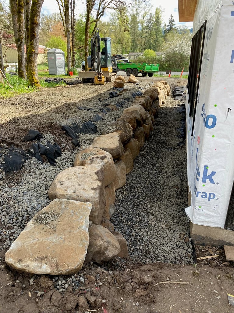 Landscaping construction site with large stone edging being installed along a gravel pathway, excavator and green equipment visible in background