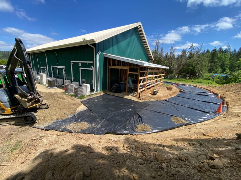 Green farmhouse with white roof under construction, black plastic ground covering, mini excavator and wooden structure visible, forest background